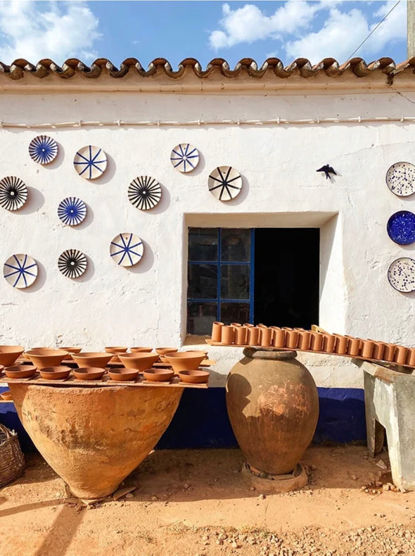 Exterior of a Portuguese house. White walls are adorned with striped ceramic plates and terracotta pots are drying in the sun in front of a blue painted window.