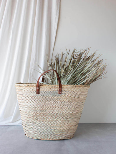 Basket bag with short tan leather handles on a grey floor and white fabric background. Palm leaves rest in the bag.