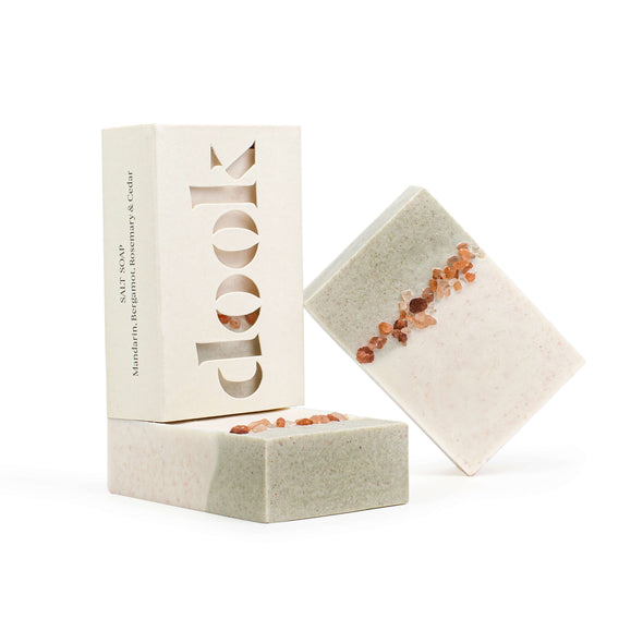 Two rectangular soaps, one lies flat and the other rests diagonally against it. Each soap is two thirds white and one third pale green, pink himalayan salt grains lie along the join between the two colours. A soap encased in its cardboard packaging sits atop the soap lying flat. The brand name 'dook' is cut out on the front of the packaging box.