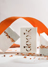 Three rectangular soap bars. Each soap is two thirds white and one third pale green, pink himalayan salt grains lie along the join between the two colours. One soap is encased in its cardboard packaging with the brand name 'dook' cut out on the front of the packaging box. Pink himalayan salt grains are scattered around the soap bars and a curled orange form provides the backdrop.
