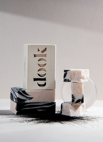 Three rectangular soaps with a white, black and cream pattern. Stacked on top of each other, the top bar sits upright and is encased in its cardboard packaging. The brand name 'dook' is cut out on the front of the packaging box. Loose charcoal fans out from the base of the pile.