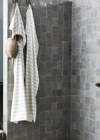 A grey tiled shower with two black metal hooks at the top. From the hooks hang two bath towels in a cream and grey stripe design and a small round basket.