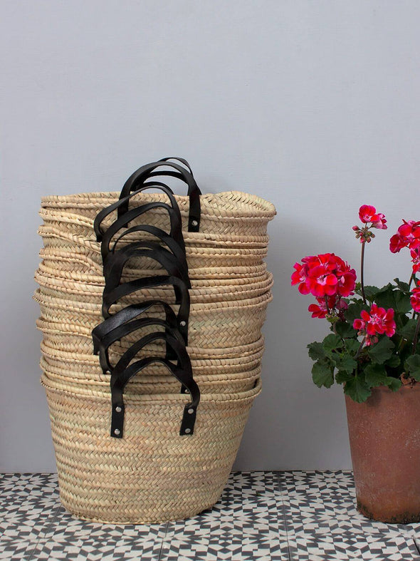 Stack of basket bags with short black leather handles sitting next to a red geranium in a terracotta pot.