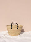 Basket bag with short black leather handles in front of a pale pink wall.