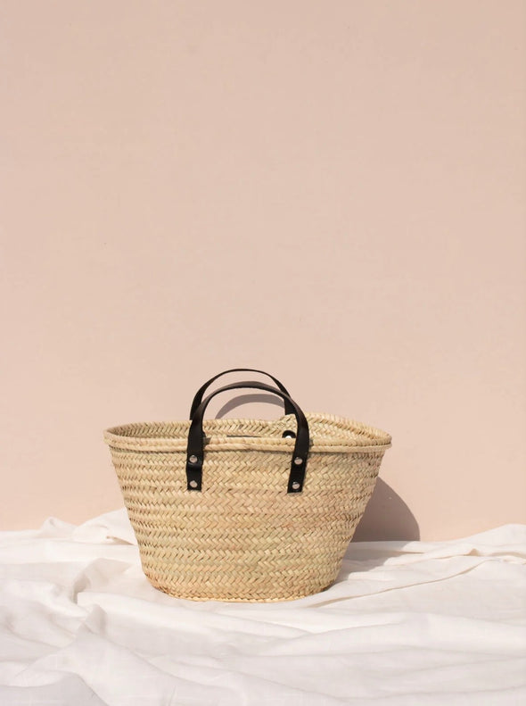 Basket bag with short black leather handles in front of a pale pink wall.