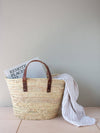 Basket bag with short tan leather straps. A hardback book and hammam towel rest inside the bag.