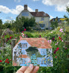 A person's hand holding up a greetings card of a house. The house has sky blue window frames and a red tiled roof. Behind the card is an real house  with lots of greenery and red and pink flowers in the foreground.