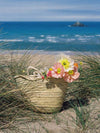 Basket bag sitting among grasses on a sandy dune in front of a blue sea. Peach and white poppies rest in the bag.