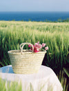 Basket bag on a table covered in a white tablecloth in front of a field of grasses and blue sea beyond. Pink flowers rest in the basket bag.