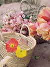 Two basket bags on a sandy beach. The bags are full of pink flowers and red and yellow poppies.