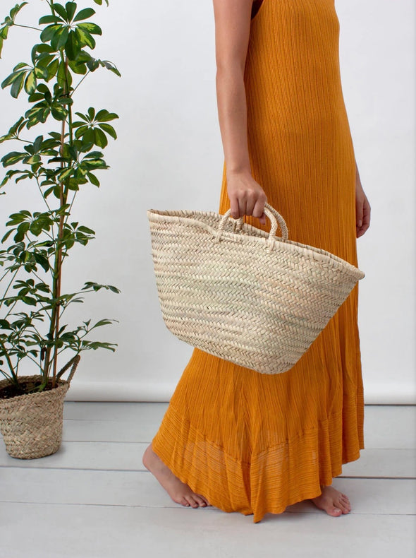 Woman in an orange dress holding a basket bag. A tall green plant sits to the side of the woman.