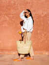 Woman standing in front of a terracotta wall holding a large basket bag with long tan leather straps.