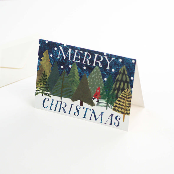 Greetings card depicting a snowy woodland scene with a person in a red cape walking through the trees. Text reads Merry Christmas.