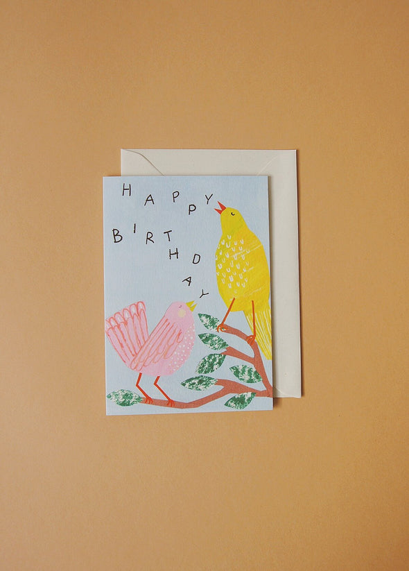 Greetings card depicting a pink bird and a yellow bird perched on a branch. Text reads happy birthday, the letters are jumbled so it looks like the birds are singing.