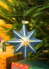 Decorative star ornament hanging on a Christmas tree with blurred gift boxes in the background.