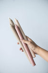 A hand holding a selection of tall dinner candles. It is a mix of dusky pink and beige candles.