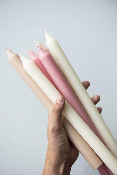 A hand holding a selection of tall dinner candles. It is a mix of pink, white and beige candles.