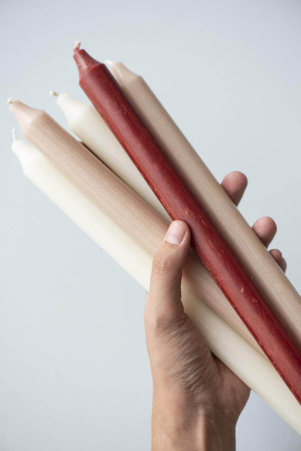 A hand holding a selection of tall dinner candles. It is a mix of red, white and beige candles.