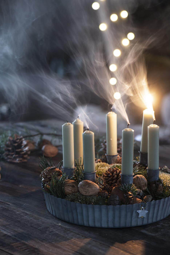 A wooden tabletop on which sits a candle display in a round metal tray. There are seven short light green dinner candles which have just been blown out except for one remaining lit candle. There is smoke coming from the unlit candles. Around the bottom of the candles are pine cones, moss and nuts.