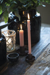 A wooden table on which sits a black glossy vase and two burning candles in short black candle holders. Both candles are a dusky pink colour, one is short and the other tall and thin.