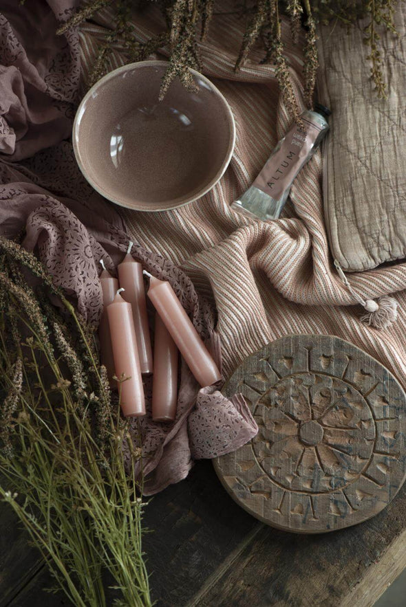 A wooden tabletop partially covered by a dusky pink cloth. Lying on top are a pink ceramic bowl, a round wooden board with carved patterns, some foliage and five short dusky pink dinner candles.