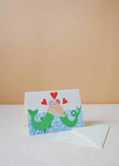 A greetings card with a painted image of two merpeople embracing in the sea. Red love hearts are dotted around their heads.