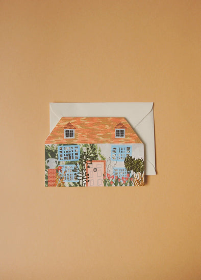 Greetings card in the shape of a house. It has a pale pink door, sky blue window frames and a red tiled roof. Lots of flowers and greenery grow infront and up the walls.