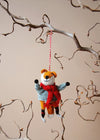 Wool fox ornament with a red scarf and wearing skates, hanging from a branch against a beige background