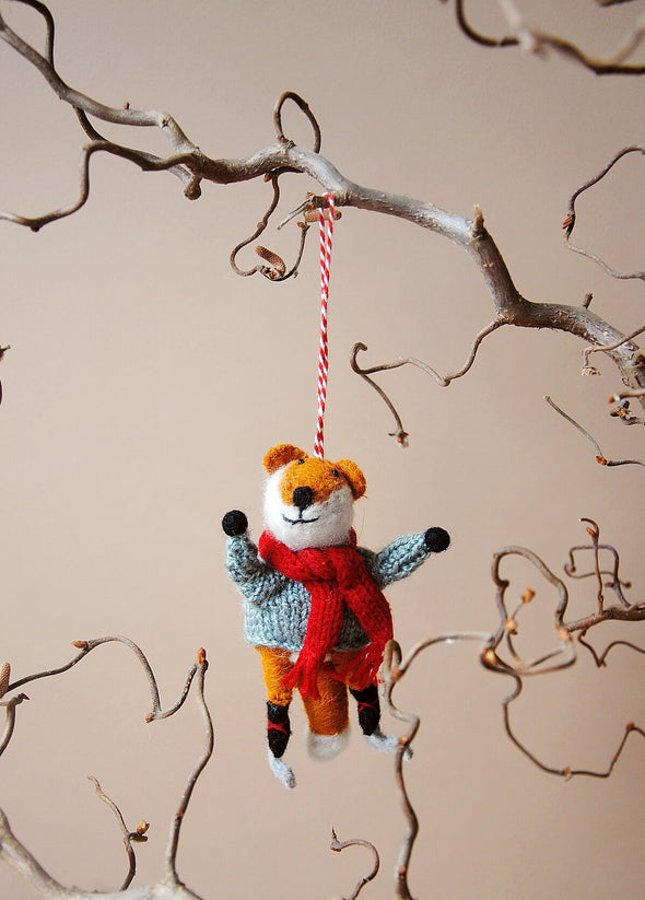 Wool fox ornament with a red scarf and wearing skates, hanging from a branch against a beige background