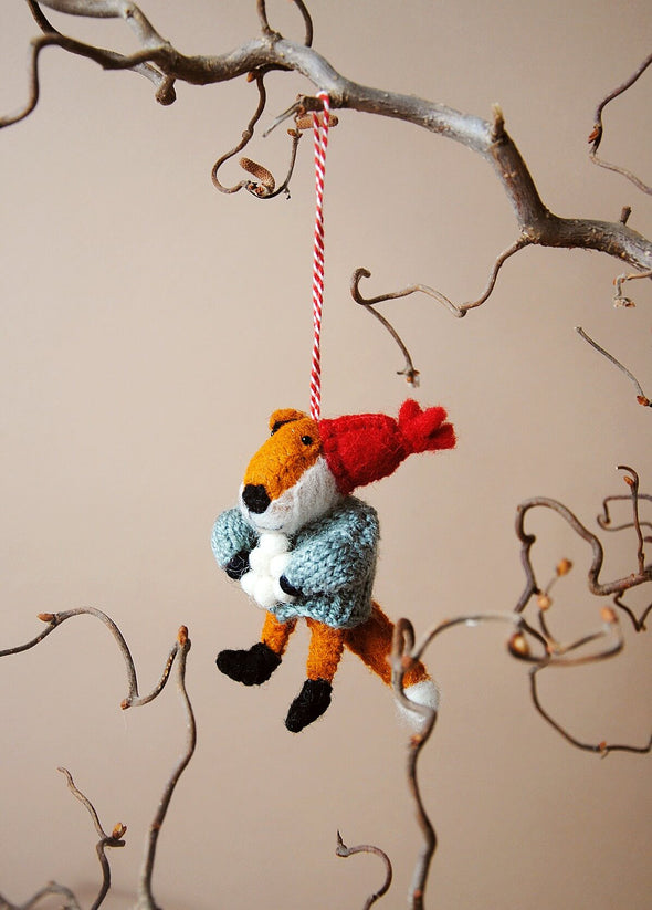 Wool fox decoration with a red hat and holding a pile of snowballs, hanging from a branch against a beige background