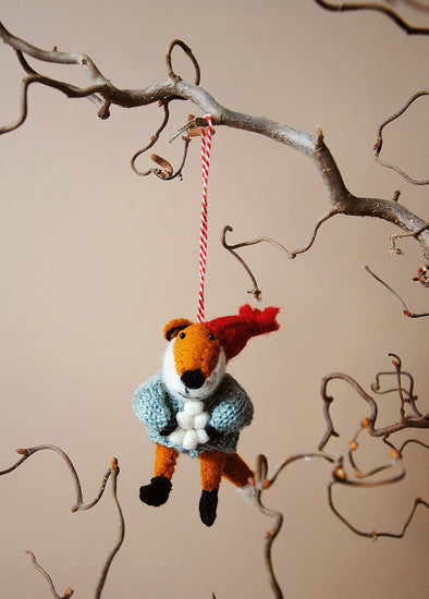 Wool fox ornament with a red hat and holding a pile of snowballs, hanging from a branch against a beige background