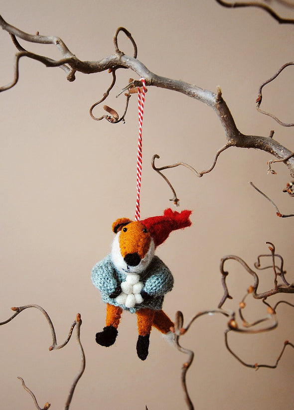 Wool fox ornament with a red hat and holding a pile of snowballs, hanging from a branch against a beige background