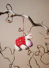 Small felted pig in a red blanket decoration hanging from a branch against a beige background