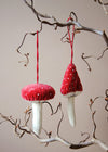 Two red and white mushroom-shaped ornaments hanging from a branch against a neutral background.