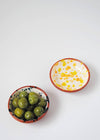 Two ceramic small bowls. One has a white glaze with blue splatter pattern on interior and the other has a white glaze with yellow and orange splatter pattern on interior. Both have a terracotta glaze on exterior. The blue bowl is filled with green olives.