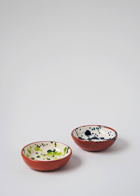 Two ceramic small bowls. One has a white glaze with blue splatter pattern on interior and the other has a white glaze with green splatter pattern on interior. Both have a terracotta glaze on exterior.
