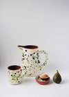 Ceramic jug, mug and small bowl. Jug and mug have a white glaze with green splatter pattern on exterior and a terracotta glaze interior. The small bowl has a white glaze with green splatter pattern interior and terracotta glaze exterior. A fig sits next to the small bowl and a cut half of fig lies in the bowl.
