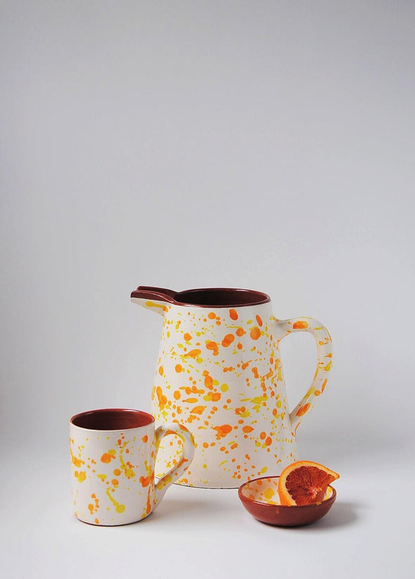 Ceramic jug, mug and small bowl. Jug and mug have a white glaze with orange and yellow splatter pattern on exterior and a terracotta glaze interior. The small bowl has a white glaze with orange and yellow splatter pattern interior and terracotta glaze exterior. A slice of blood orange lies in the bowl.