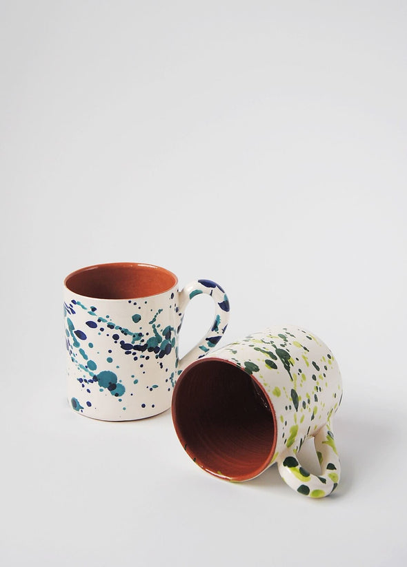 Two ceramic mugs with handles. One mug is standing upright and has a white glaze with blue splatter pattern on exterior, terracotta glaze on interior. Second mug is lying on its side and has a white glaze with green splatter pattern on exterior, terracotta glaze on interior.