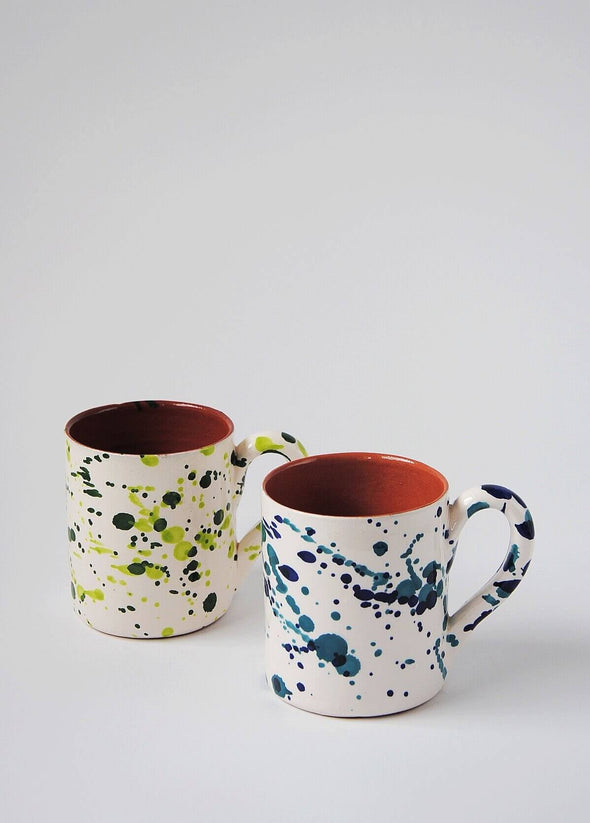 Two ceramic mugs with handles. One mug has a white glaze with blue splatter pattern on exterior. Second mug has a white glaze with green splatter pattern on exterior. Both have a terracotta glaze on interior.