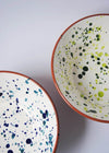 Close up of two ceramic salad bowls from above. One has a white glaze with blue splatter pattern on interior and the other a white glaze with green splatter pattern on interior. Both have a terracotta glaze on exterior.