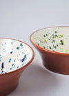 Close up of two ceramic salad bowls. One has a white glaze with blue splatter pattern on interior and the other a white glaze with green splatter pattern on interior. Both have a terracotta glaze on exterior.