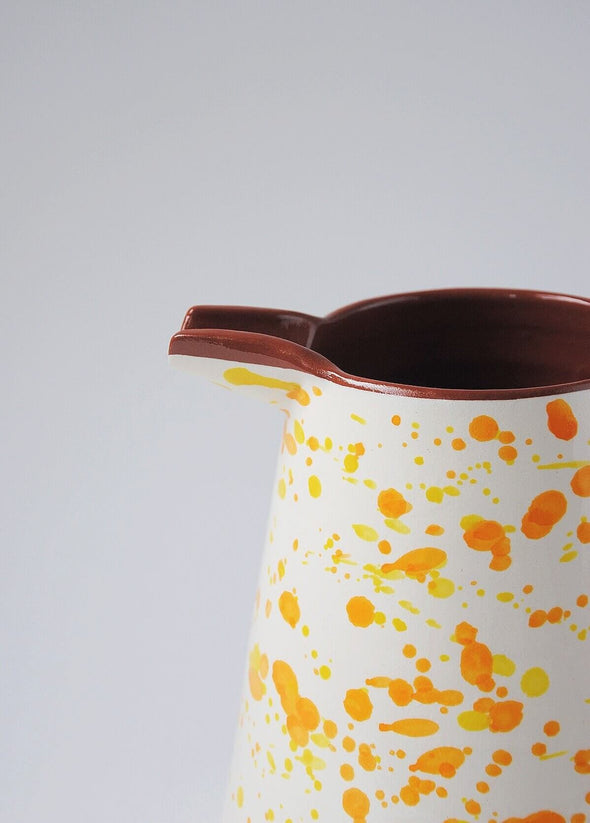 Close up of ceramic large jug. White glaze with orange and yellow splatter pattern on exterior, terracotta glaze on interior.