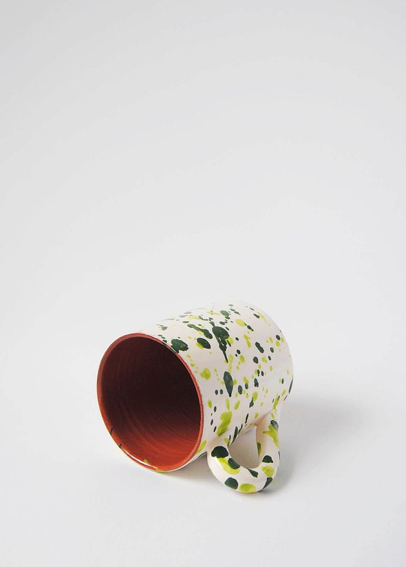 Ceramic mug with handle, lying on its side. White glaze with green splatter pattern on exterior, terracotta glaze on interior.