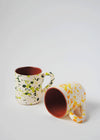 Two ceramic mugs with handles. One mug is standing upright and has a white glaze with green splatter pattern on exterior, terracotta glaze on interior. Second mug is lying on its side and has a white glaze with orange and yellow splatter pattern on exterior, terracotta glaze on interior.