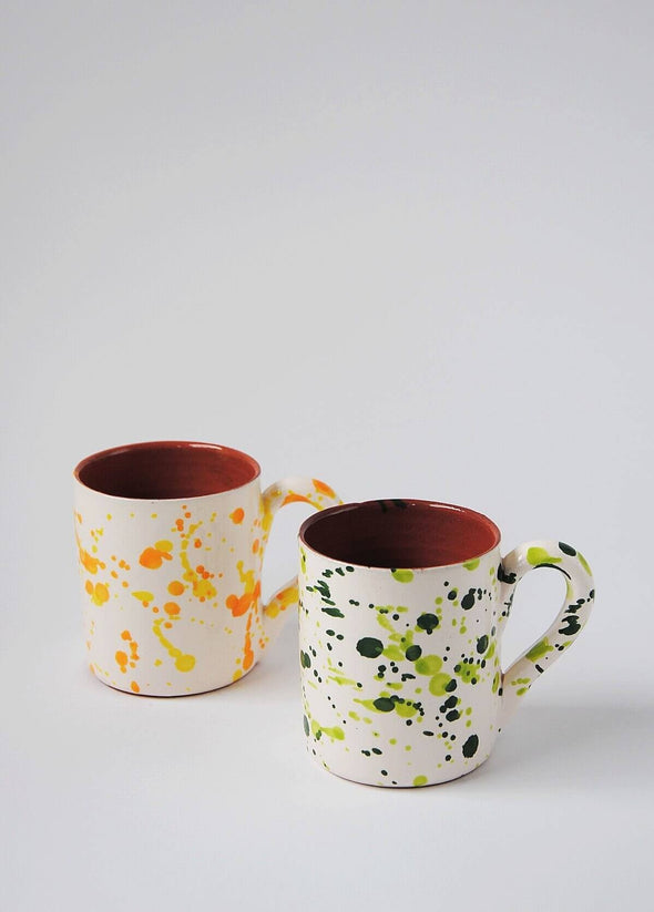 Two ceramic mugs with handles. One mug has a white glaze with orange and yellow splatter pattern on exterior. Second mug has a white glaze with green splatter pattern on exterior. Both have a terracotta glaze on interior.