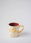 Ceramic mug with handle. White glaze with orange and yellow splatter pattern on exterior, terracotta glaze on interior.