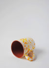 Ceramic mug with handle, lying on its side. White glaze with orange and yellow splatter pattern on exterior, terracotta glaze on interior.