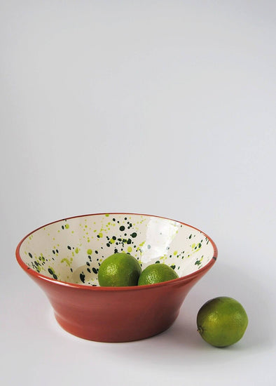 Ceramic salad bowl. White glaze with green splatter pattern on interior and terracotta glaze on exterior. Two limes rest inside the bowl and one lies next to it.
