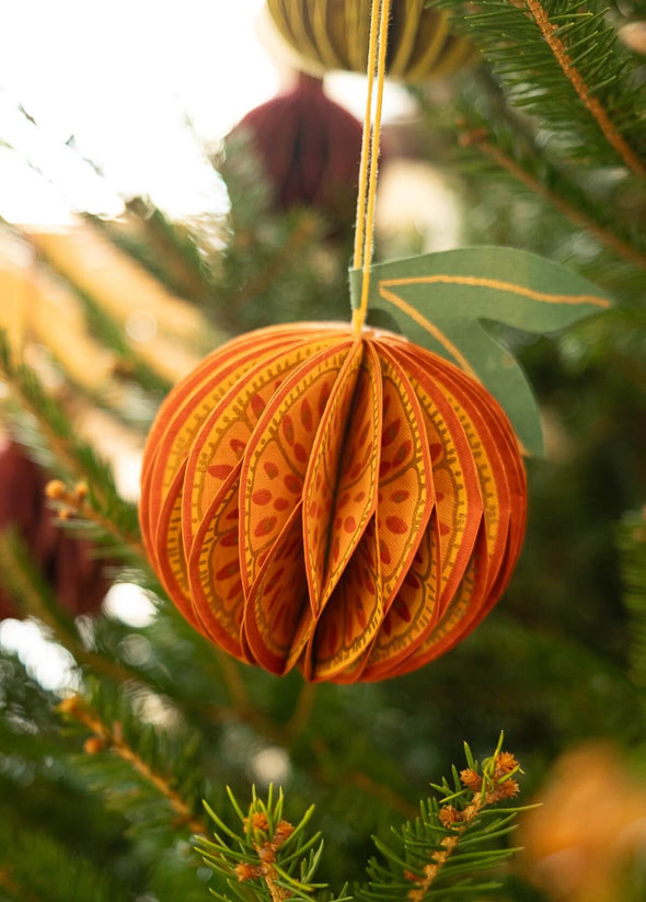 Paper honeycomb decoration in the shape of an orange. It is hanging from a Christmas tree.